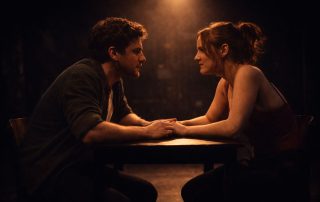 Two actors seated closely at a table under a spotlight, sharing an intense and intimate moment on stage