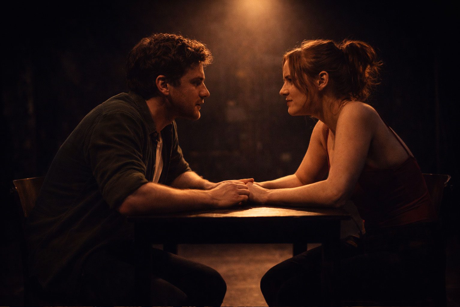 Two actors seated closely at a table under a spotlight, sharing an intense and intimate moment on stage