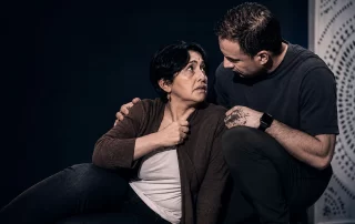 Cinematic intimate theatre scene from Constellations featuring a man comforting a distressed woman on stage under dramatic warm lighting, emphasizing emotional vulnerability and human connection in a minimalist black-box theatre setting.
