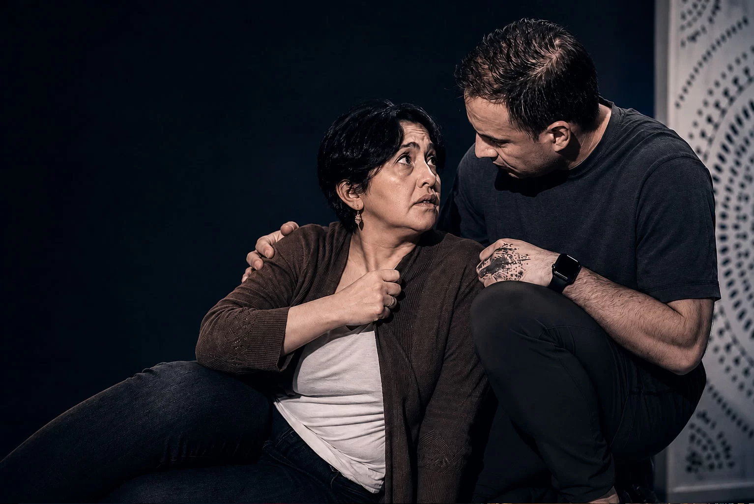 Cinematic intimate theatre scene from Constellations featuring a man comforting a distressed woman on stage under dramatic warm lighting, emphasizing emotional vulnerability and human connection in a minimalist black-box theatre setting.